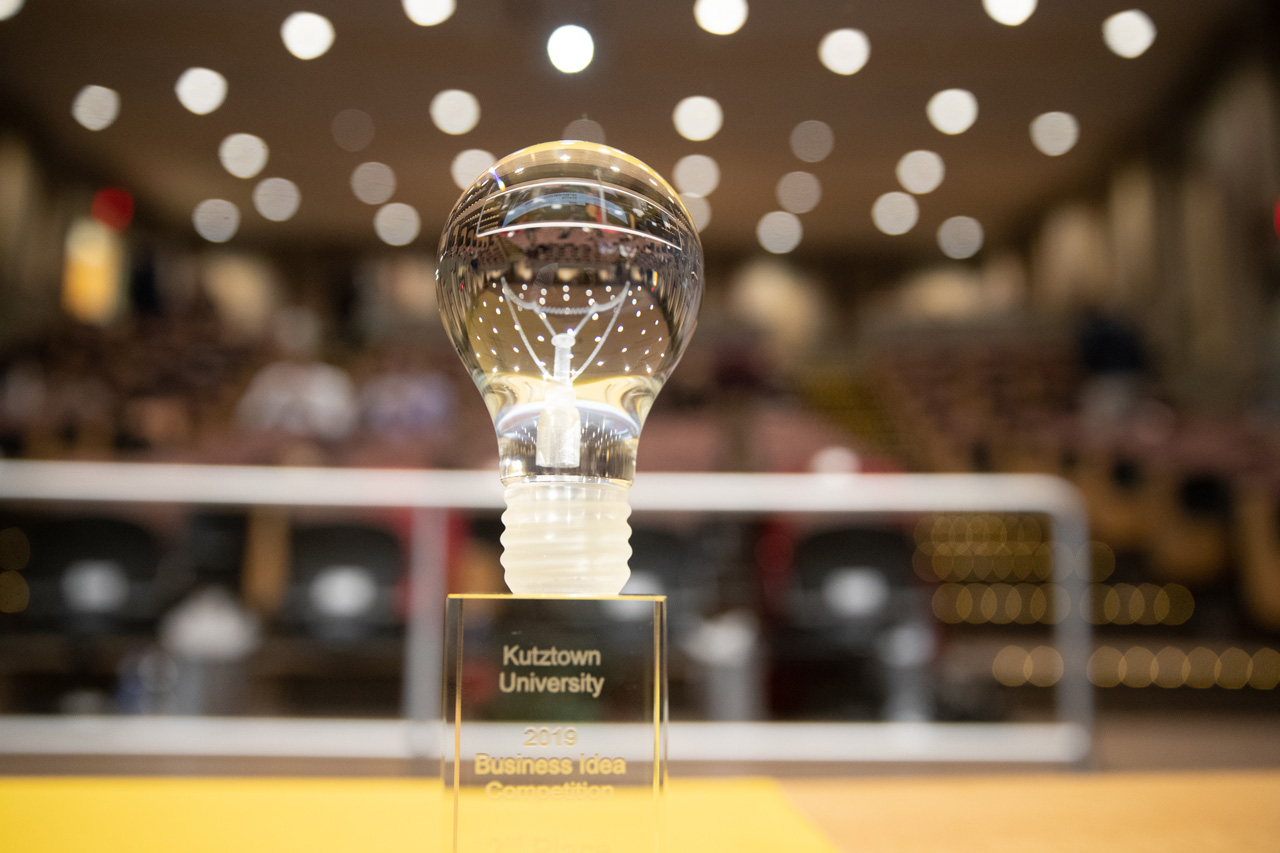 A Clear Glass lightbulb trophy that reads "Kutztown University 2019 Business Idea Competition" in the foreground of an empty auditorium