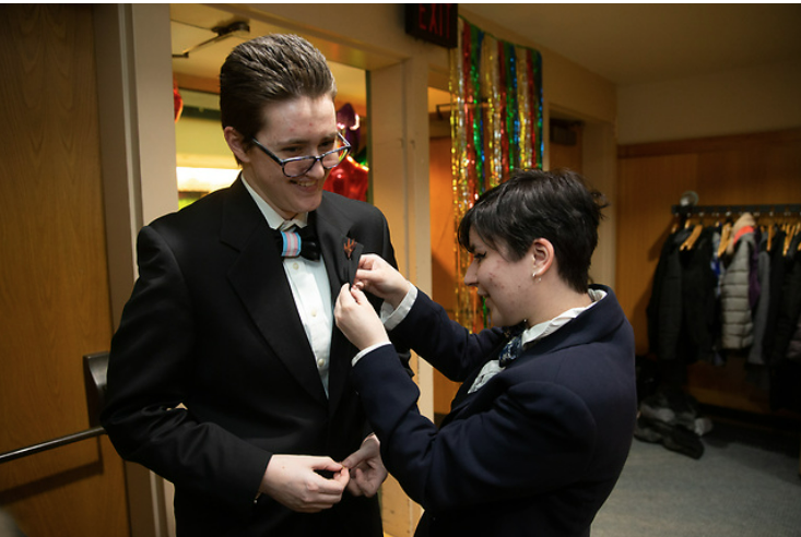 two students in tuxedos, one pinning the other indoors at pride prom