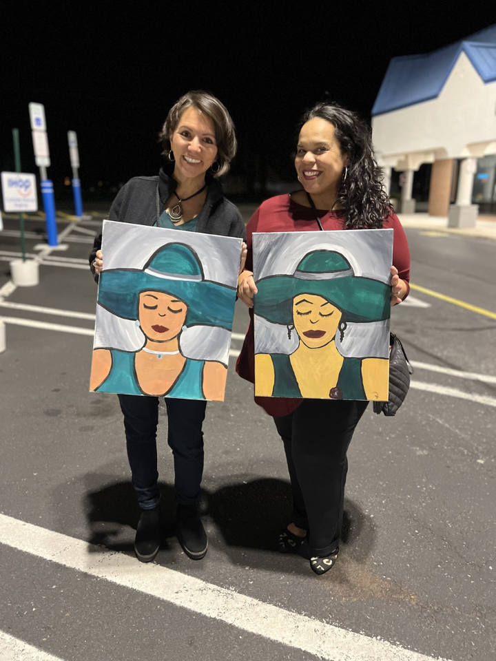 two women smiling standing outside holding paintings