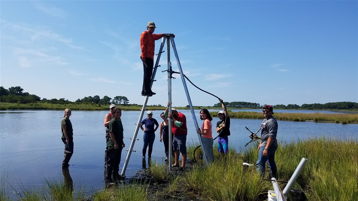 Students standing in a group around a tall tripod, which one student is standing on, in front of a lake- vibracoring in Swan Cove Pool on Assateague Island