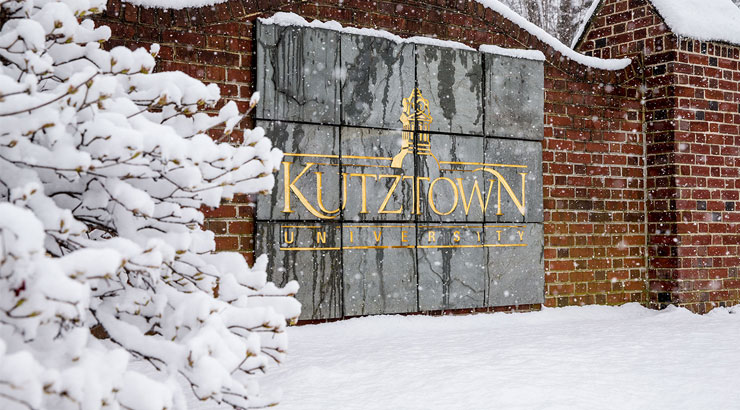 Snow falling in front of KU sign with sow on the ground and trees.