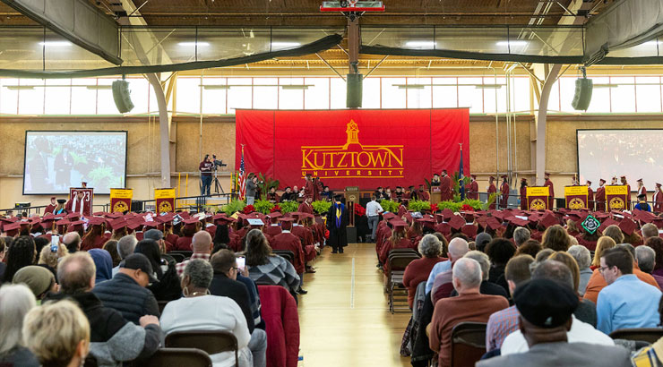 commencement ceremony as seen from the back of the field house, audience members and graduates look on as the graduates walk across the stage to receive their diploma.