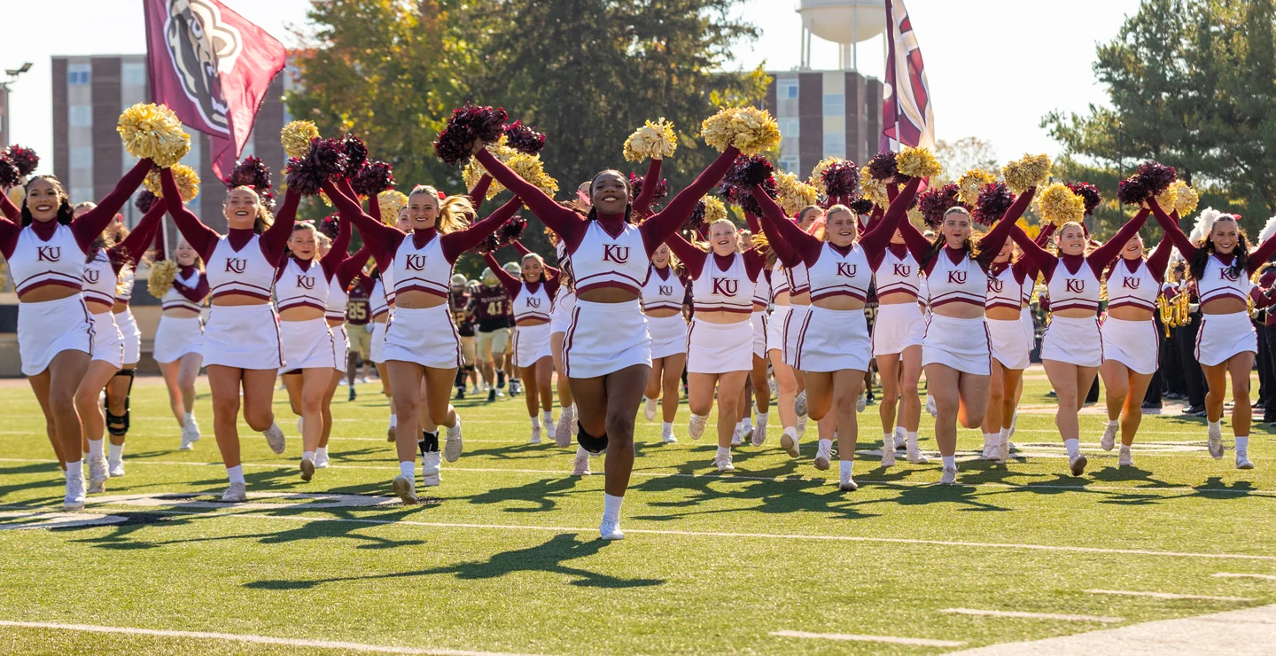 A group of diverse female Kutztown University cheerleaders running onto the football field cheering and smiling