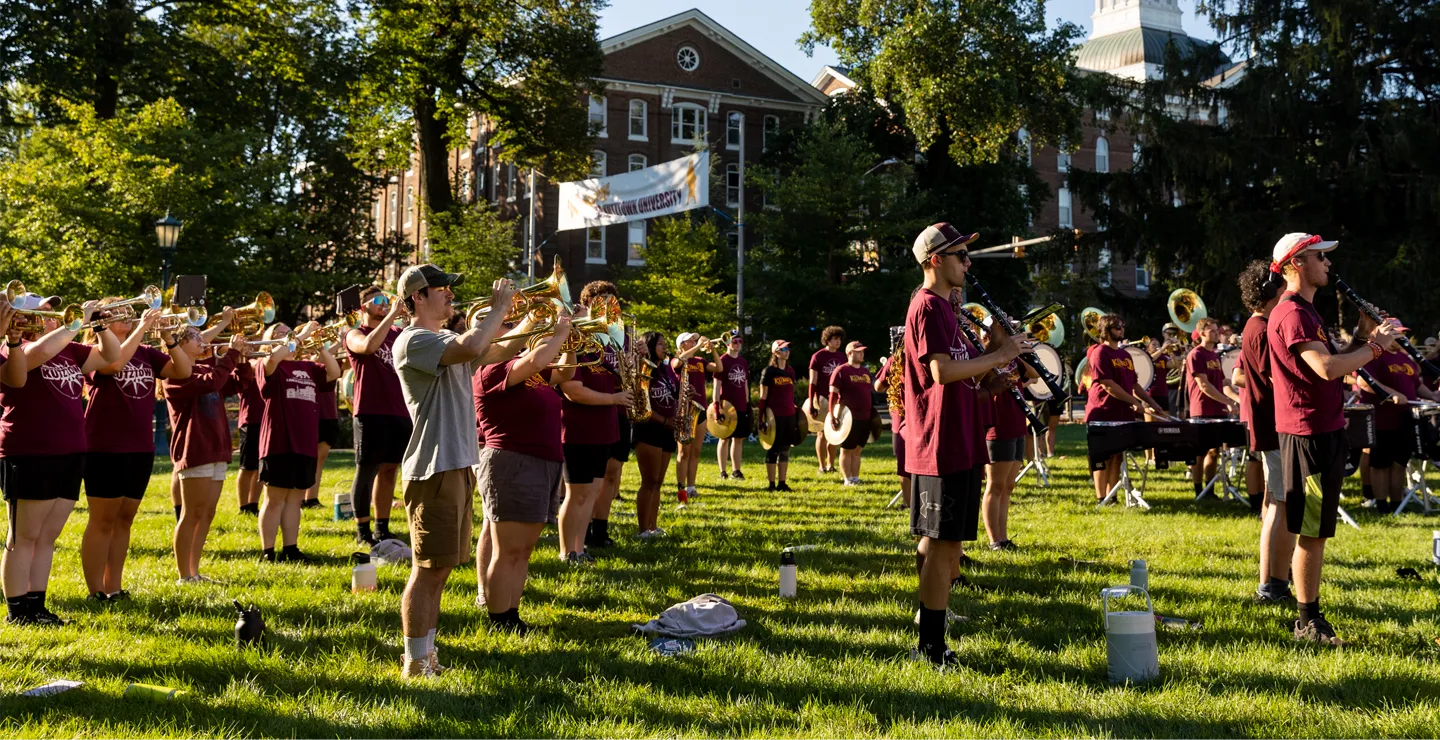 A group of male and female student in the Kutztown University marching band standing on a lawn playing instruments