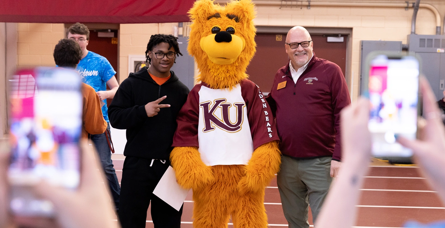 Avalanche the golden bear mascot standing with President Cavalier and an african american male student at accepted student day while others take photos of the three on cell phones