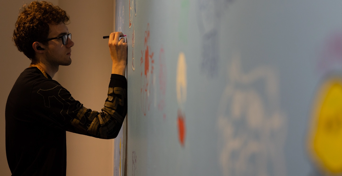 A white male student standing a drawing on an art piece that is mounted on a wall