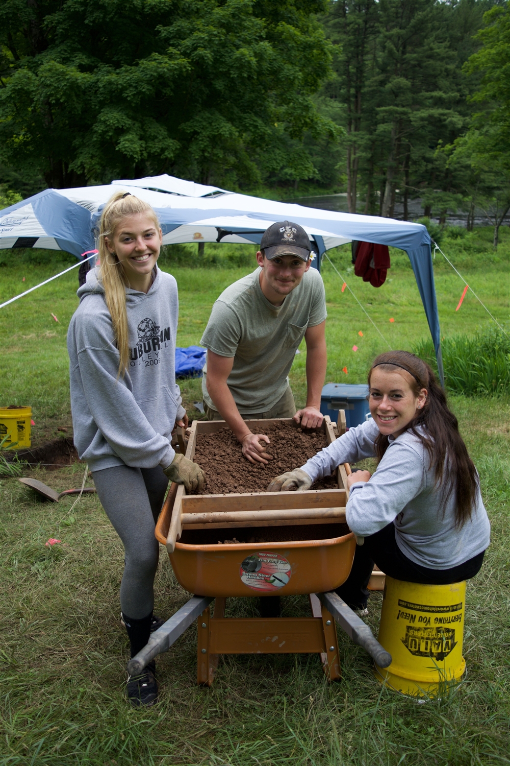 Three students standing around a wheelbarrow full of dirt, sticking their hands in and smiling at the camera 