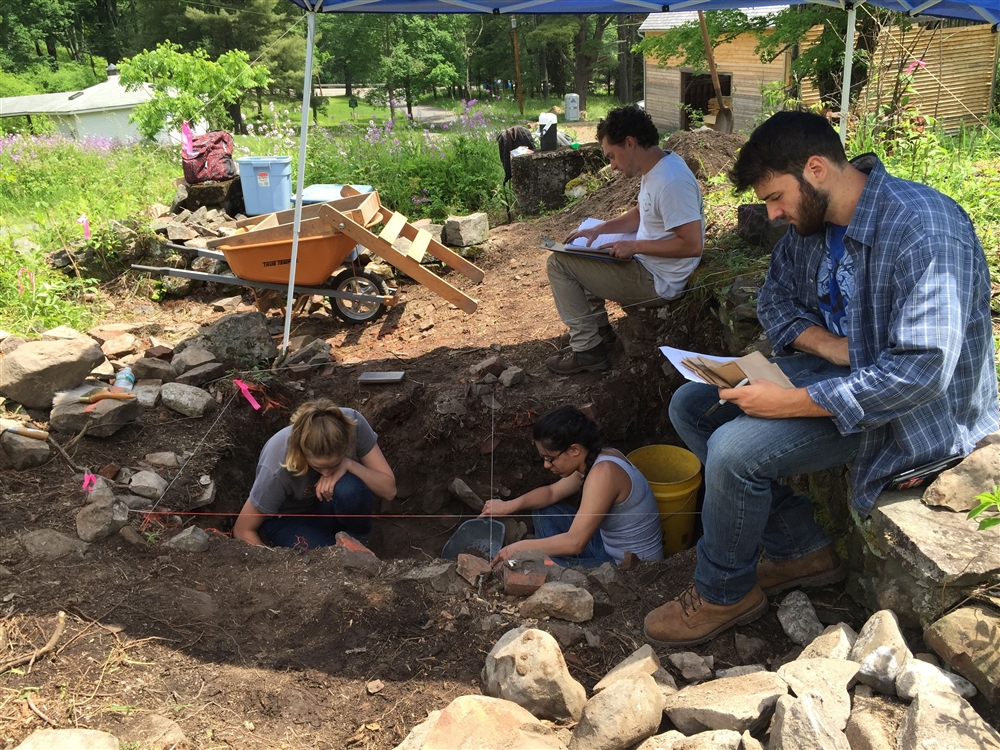 Two female students gather specimen from a dig site while two male students take notes on the ground above