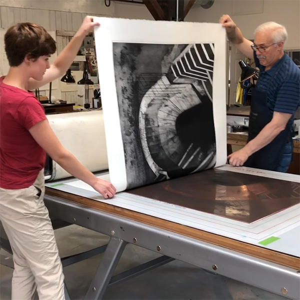 A professor and a student pull a large etching print off of a copper plate to reveal a black and white etching