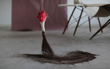 A sculpture that consists of a red face connected to a hand broom that is sweeping up charcoal-colored powder