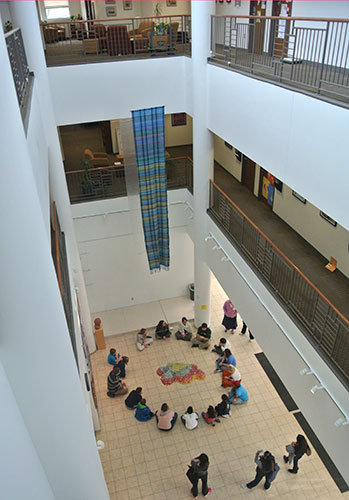 Overhead shot of students, sitting in a circle around a painting on the tile floor, taken from a balcony several floors up 