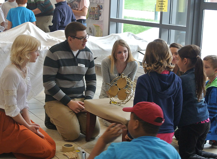 Art educators on one side of a table, showing a wooden sculpture to elementary school students on the other side of the table