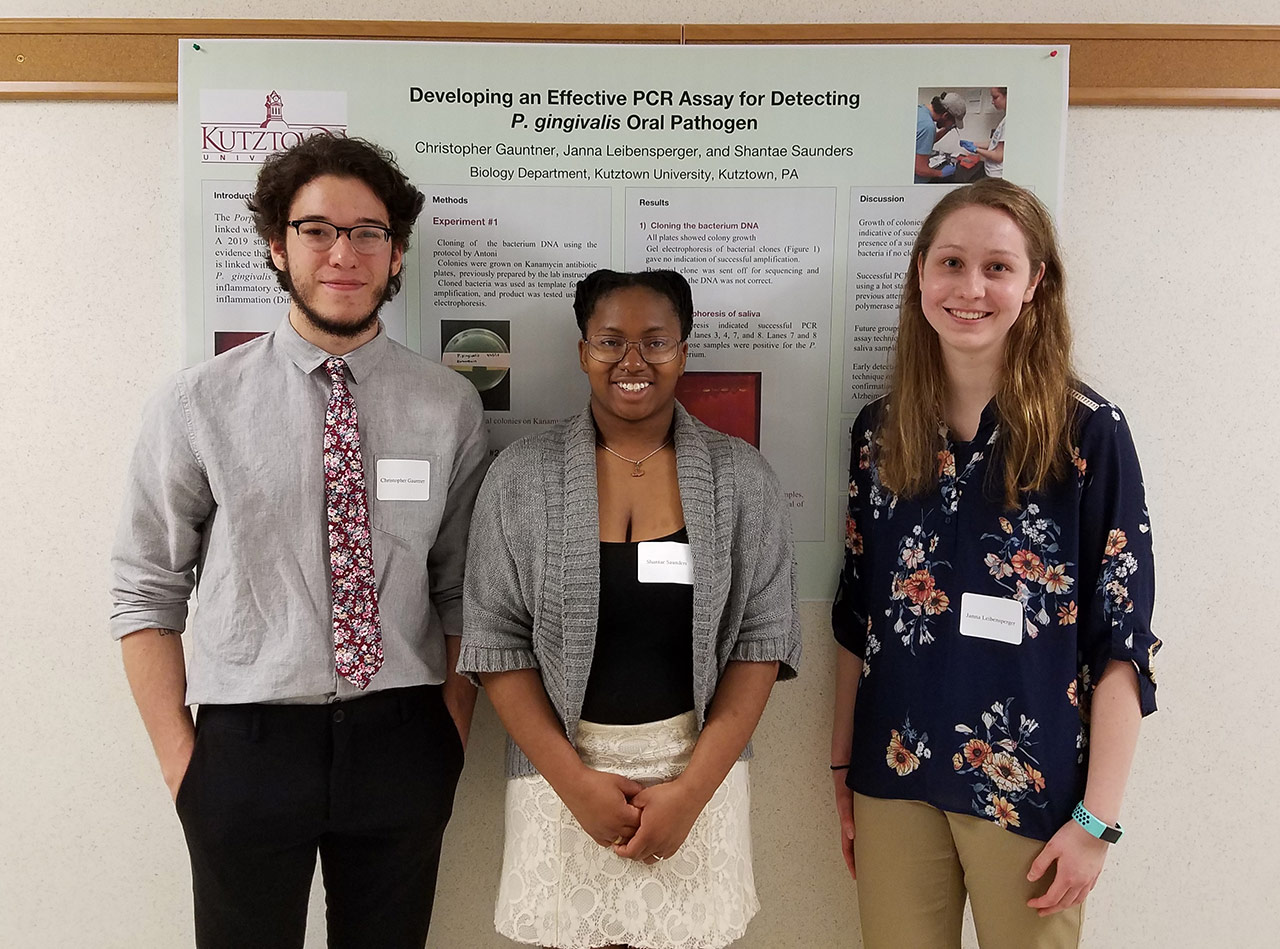 Three students standing in front of a poster on "Developing an effective PCR assay for detecting P. gingivalis oral pathogen." 