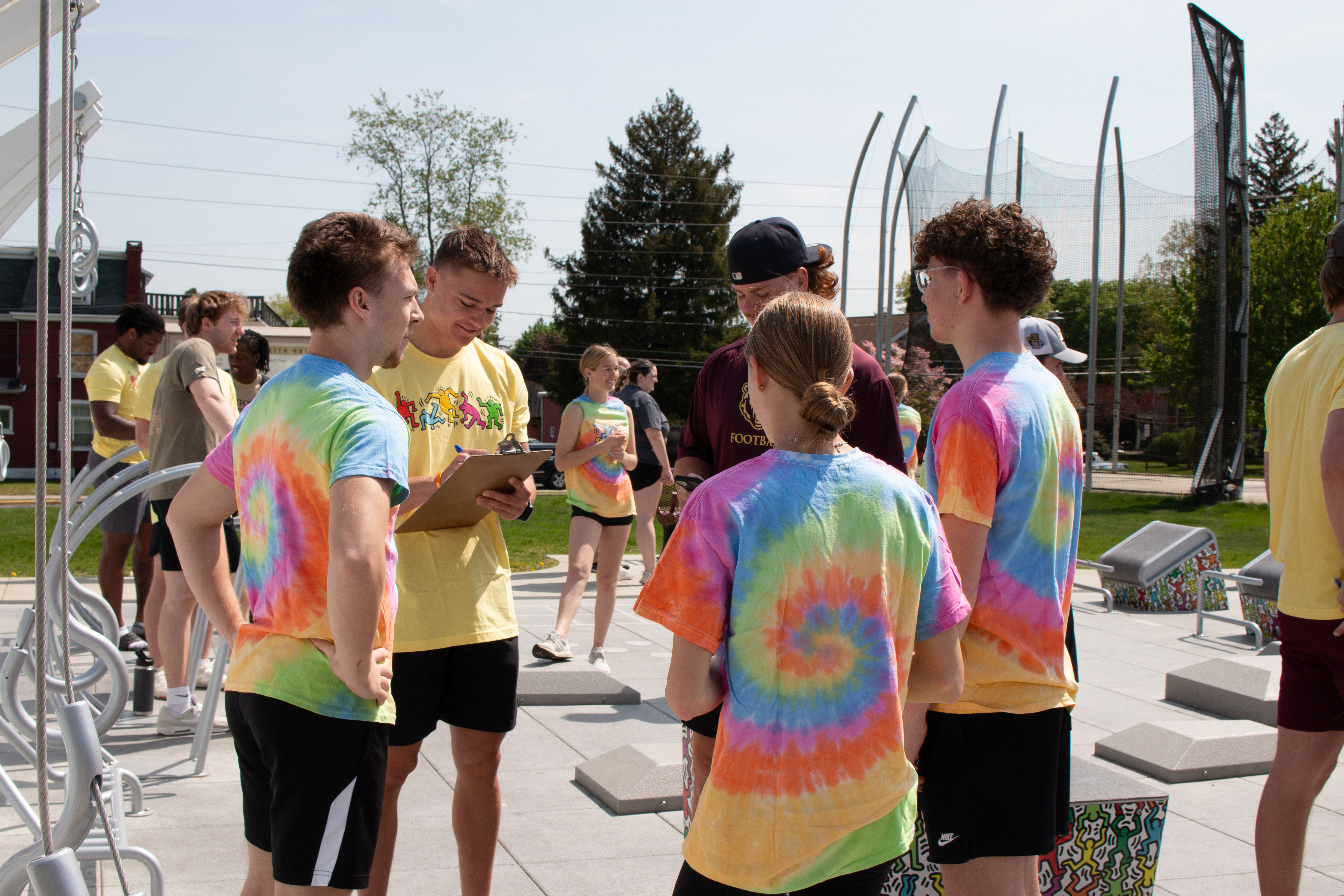 Students wearing colorful tie-dye shirts gather at Kutztown University's Keith Haring Fitness Court, discussing activities and reviewing a clipboard during the annual Fitness Clinic and Competition.