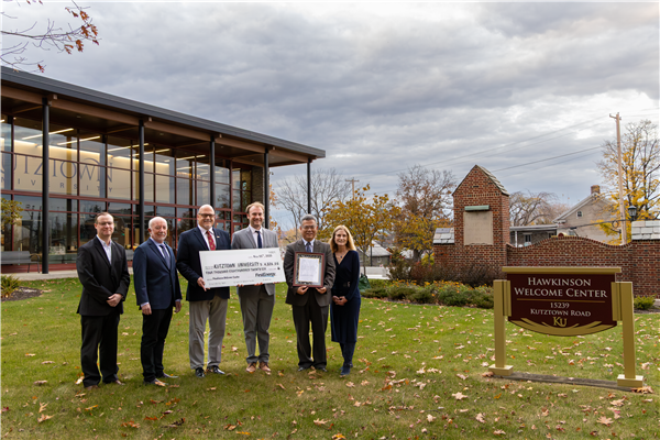 Six people stand outside Kutztown University’s Hawkinson Welcome Center holding an oversized check and framed certificate. The large check from FirstEnergy is made out to Kutztown University for $4,336.40. The group poses in front of the building and sign that reads “Hawkinson Welcome Center, 15239 Kutztown Road, KU,” with autumn leaves scattered on the grass and a cloudy sky above.