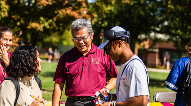Dr. Chao smiles while interacting with students outdoors on campus during a sunny fall day.