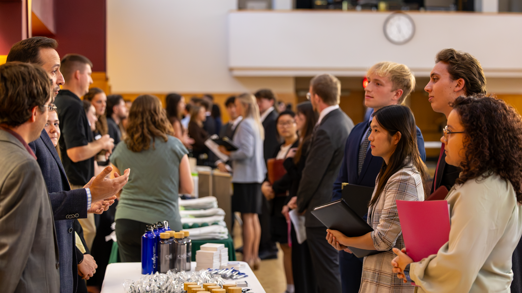 Students engage with employers at a College of Business Meet the Firms networking event. Business professionals speak with students dressed in professional attire, while other attendees network throughout the crowded event space.