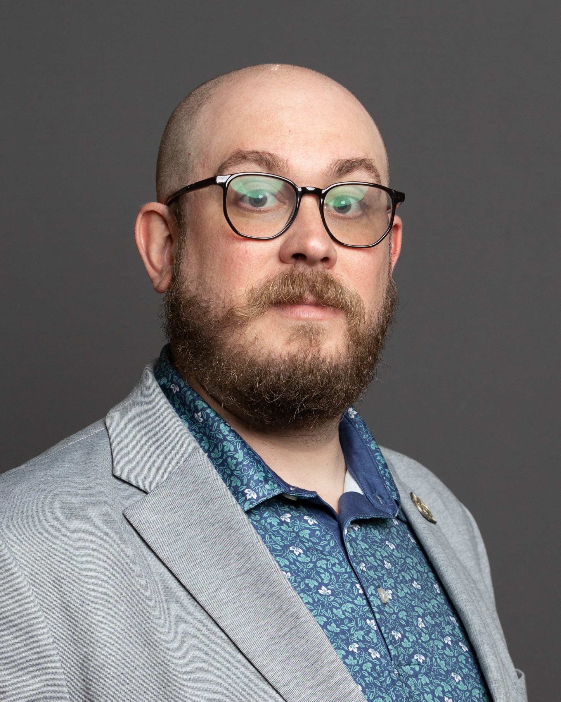 A white man with glasses and a beard is seated at a desk wearing a burgundy polo and a brown suit jacket