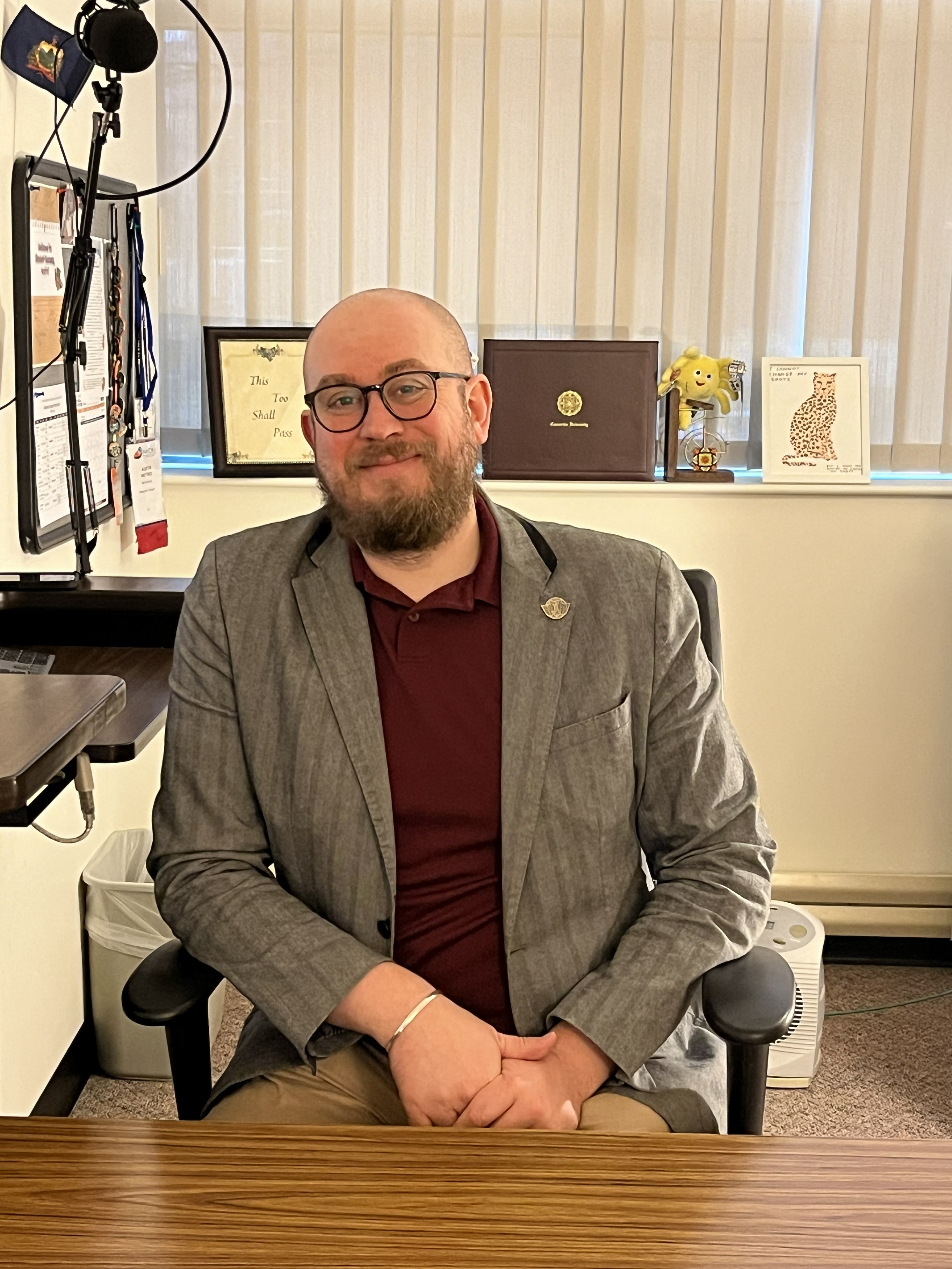A white man with glasses and a beard is seated at a desk wearing a burgundy polo and a brown suit jacket