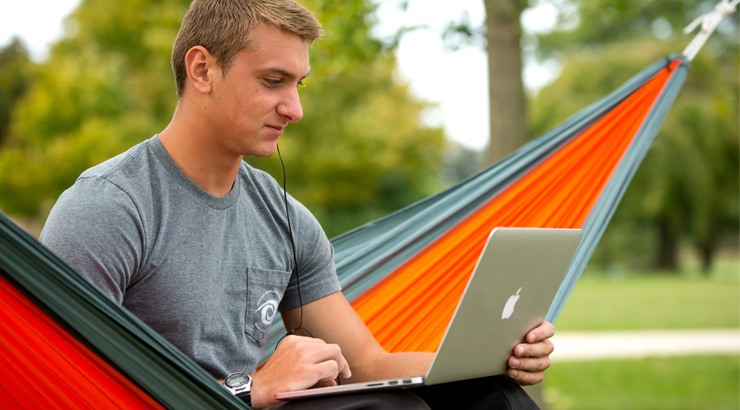 Young caucasian male sitting in a hammock outside working on a laptop set on his lap