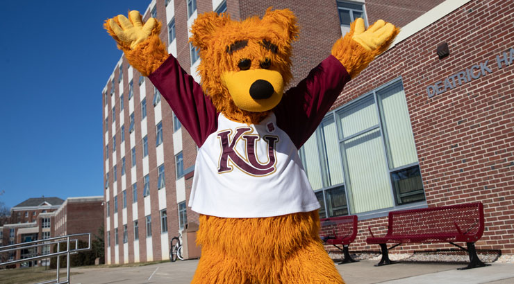 Avalanche the mascot standing in front of a residence a hall with his arms up on a sunny day in a celebratory pose