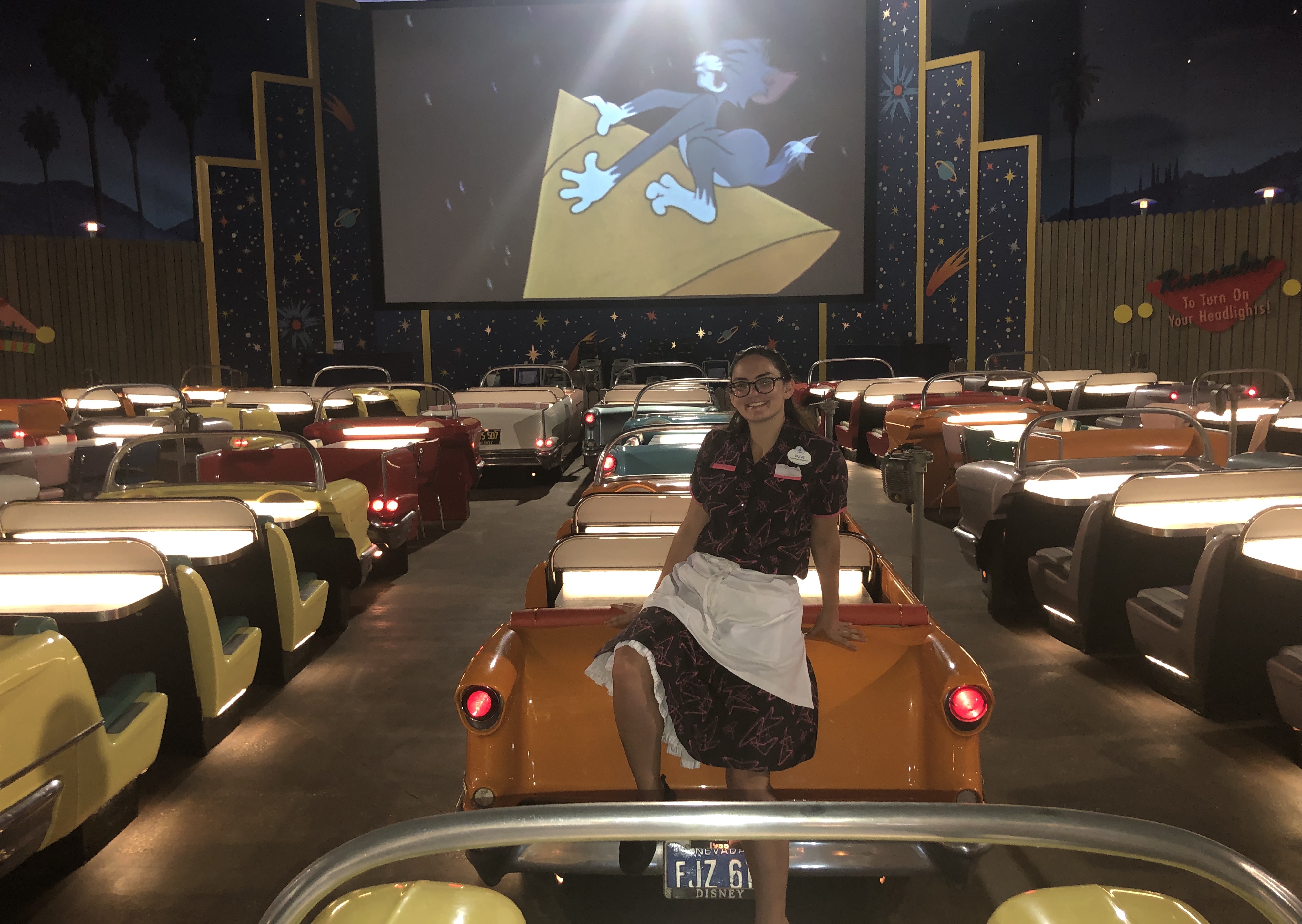 Olivia Bauer, wearing a costume and apron and sitting on the hood of a prop car, surrounded by diner booths with a screen showing a Tom and Jerry episode in the background 