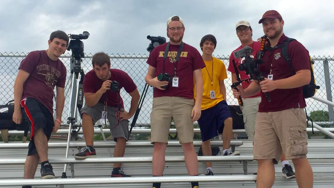 Male media students smiling on the bleachers of Andre Reed stadium with their filming equipment 