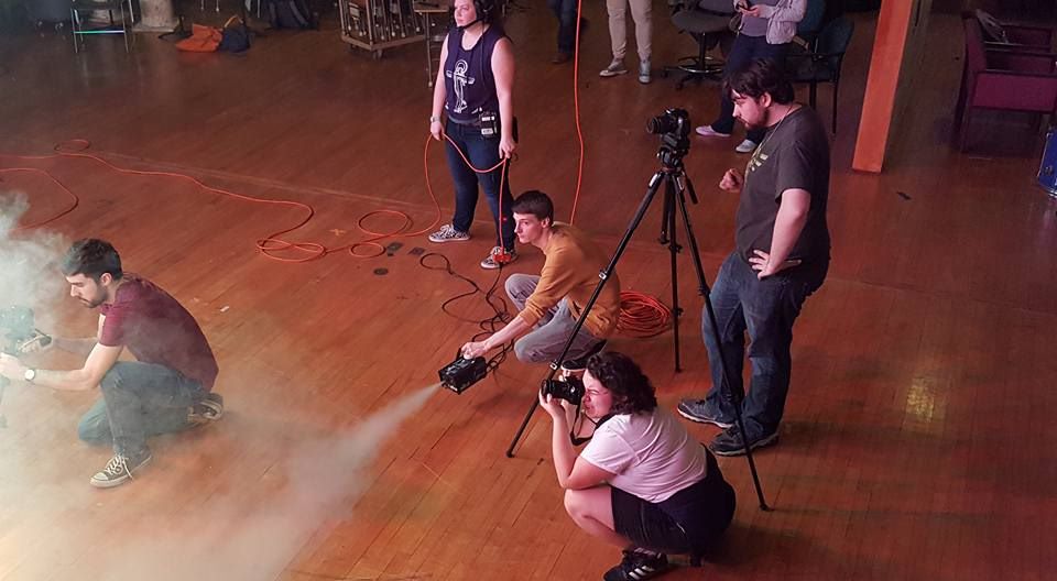 Performing arts students shooting a scene, with one behind the camera and two crouching with fog machines. 