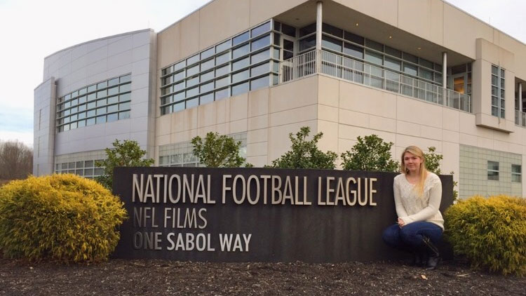 Female student kneeling next to the welcome sign at NFL films studio, with the studio building in the background.