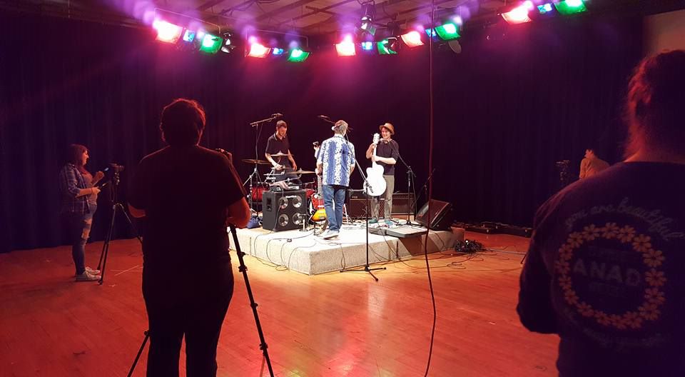 Small band standing on stage and setting up for a performance in the tv studio control room, surrounded by KU tv camera crew 
