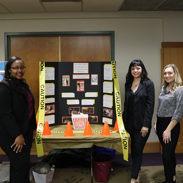 Three students standing on either side in front of a poster presentation with caution tape, traffic cones and popcorn on the table in front 