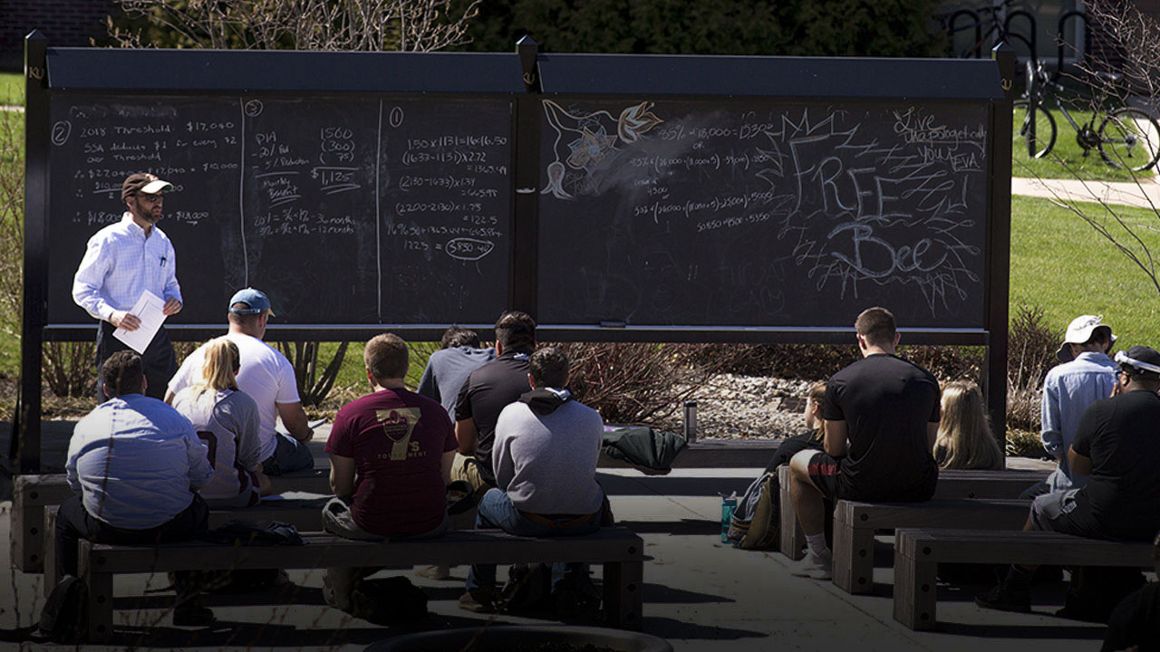 Professor lecturing to a full outside classroom with a blackboard with mathematic symbols 