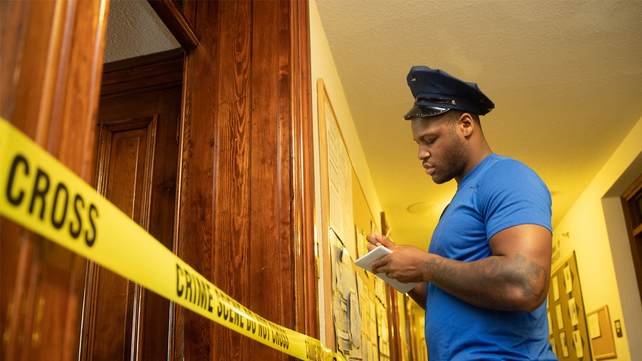 Student in a police cap taking notes at mock crime scene surrounded by tape that reads "crime scene, do not cross."