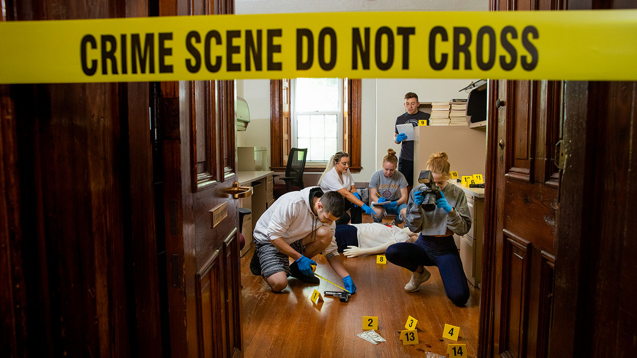 Crime scene scenario with a group of students analyzing the room for clues. Yellow tape reads, "Crime Scene Do Not Cross"