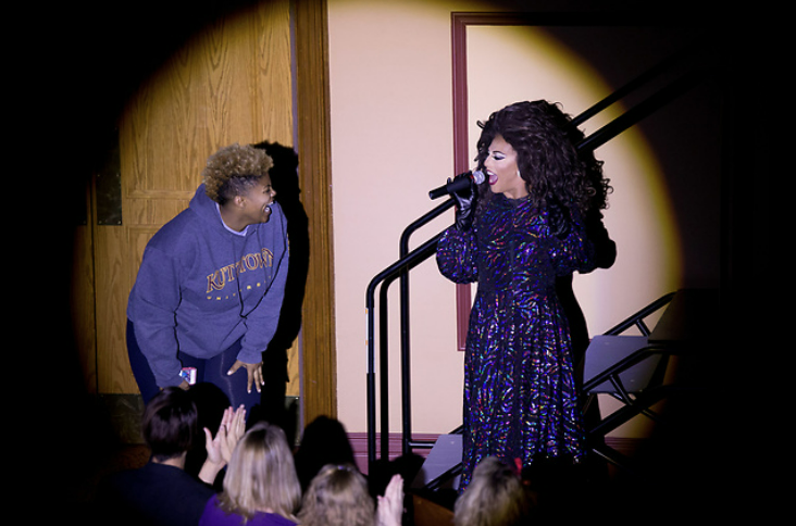 a student and performing drag queen facing each other on stage in spotlight