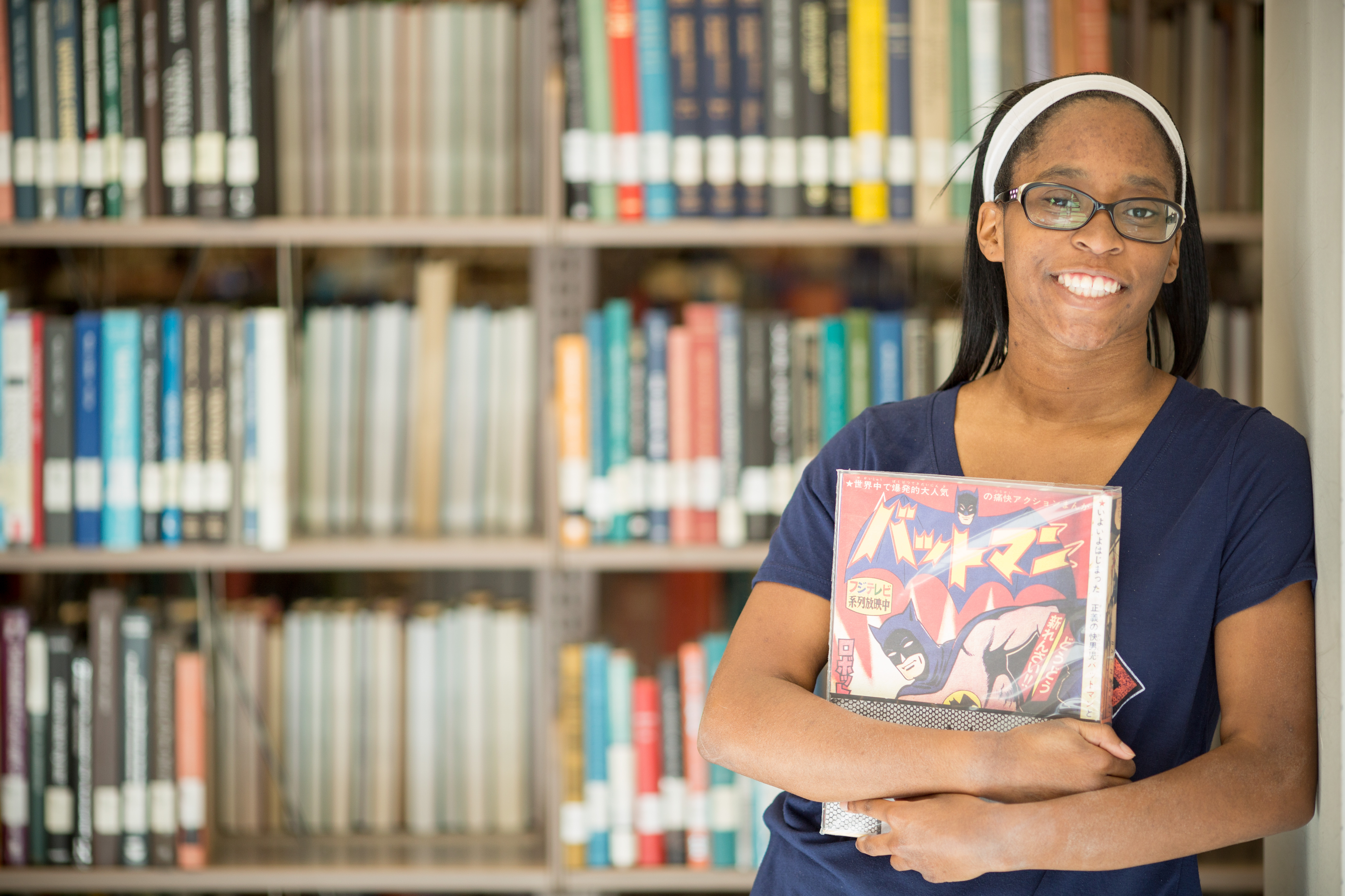 student smiling in the Rohrbach Library, holding a comic book and standing in front of a book shelf