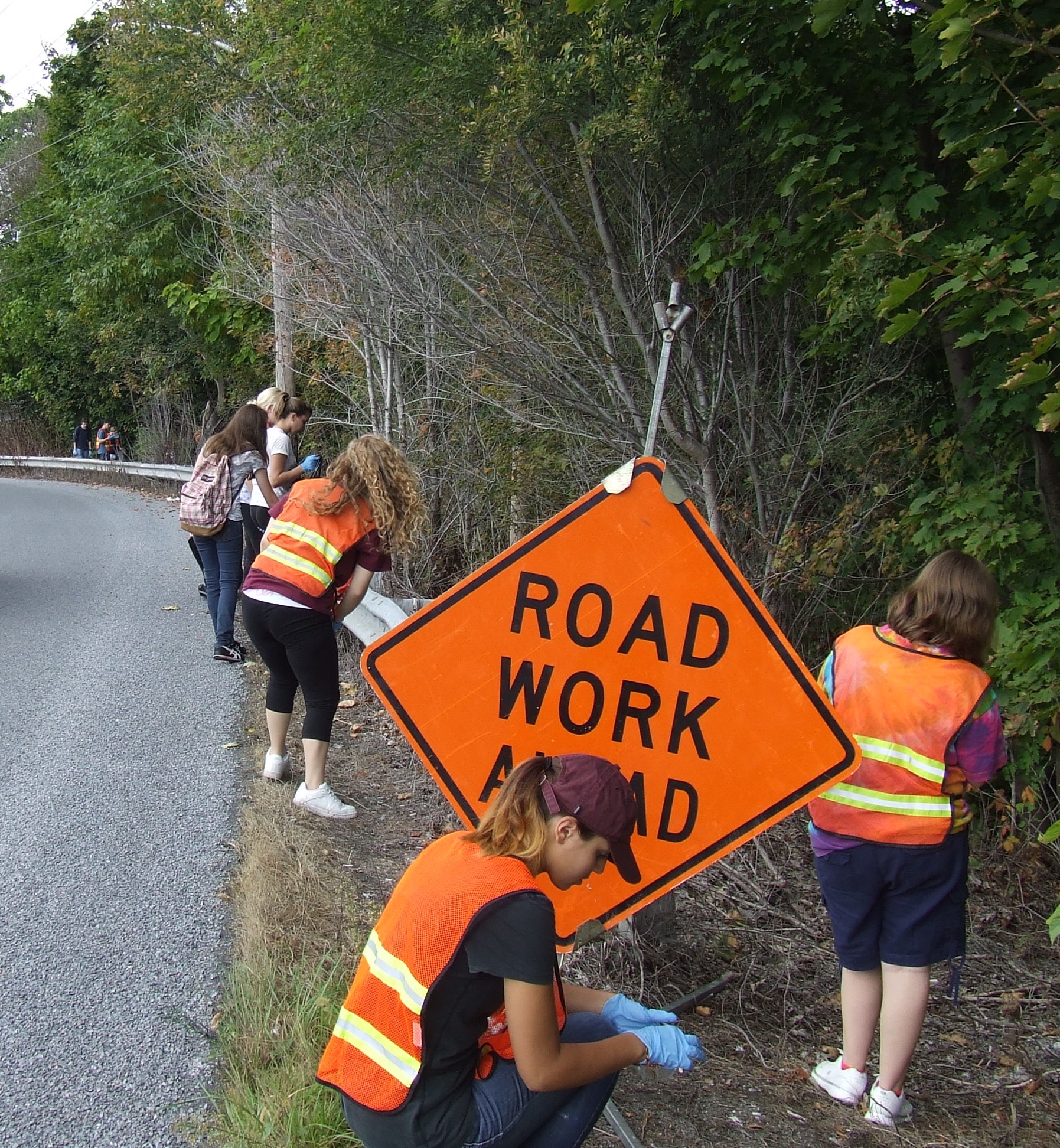 students in orange traffic vests, picking up trash along a road next to an orange sign that reads "road work ahead."