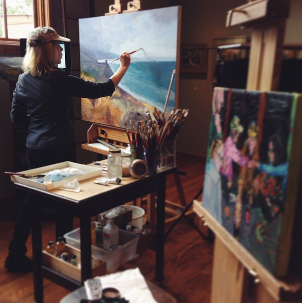 Female student painting a beach on canvas, with another painting of three women holding hands in the foreground