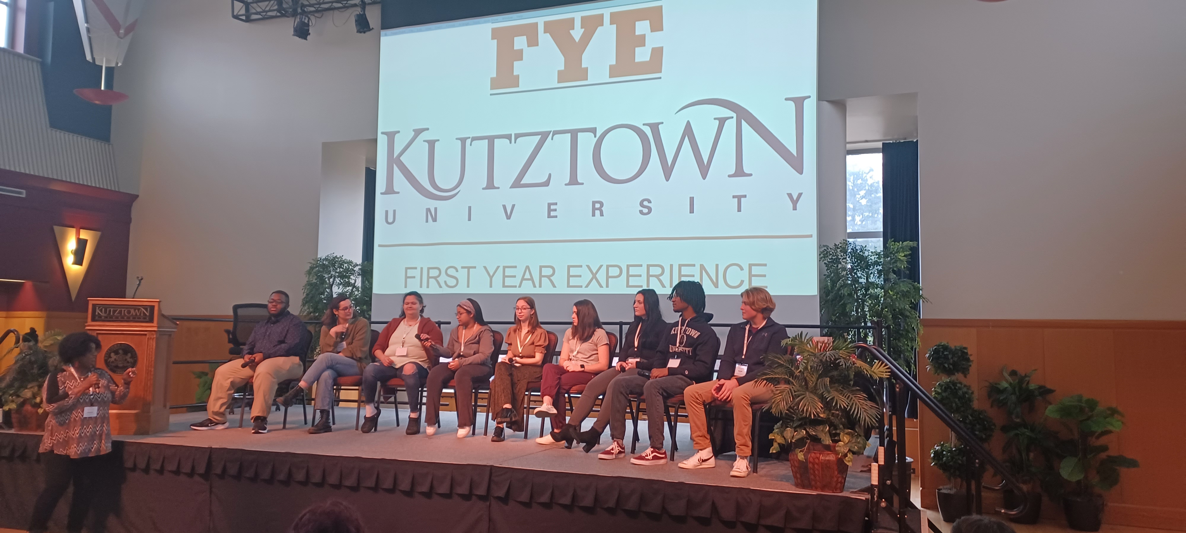 A lineup of KU students seated next to each other on stage in front of a slideshow presentation with the Kutztown University First Year Experience logo 