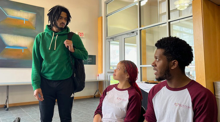 male student standing, two students, one male one female, sitting, at debate event.