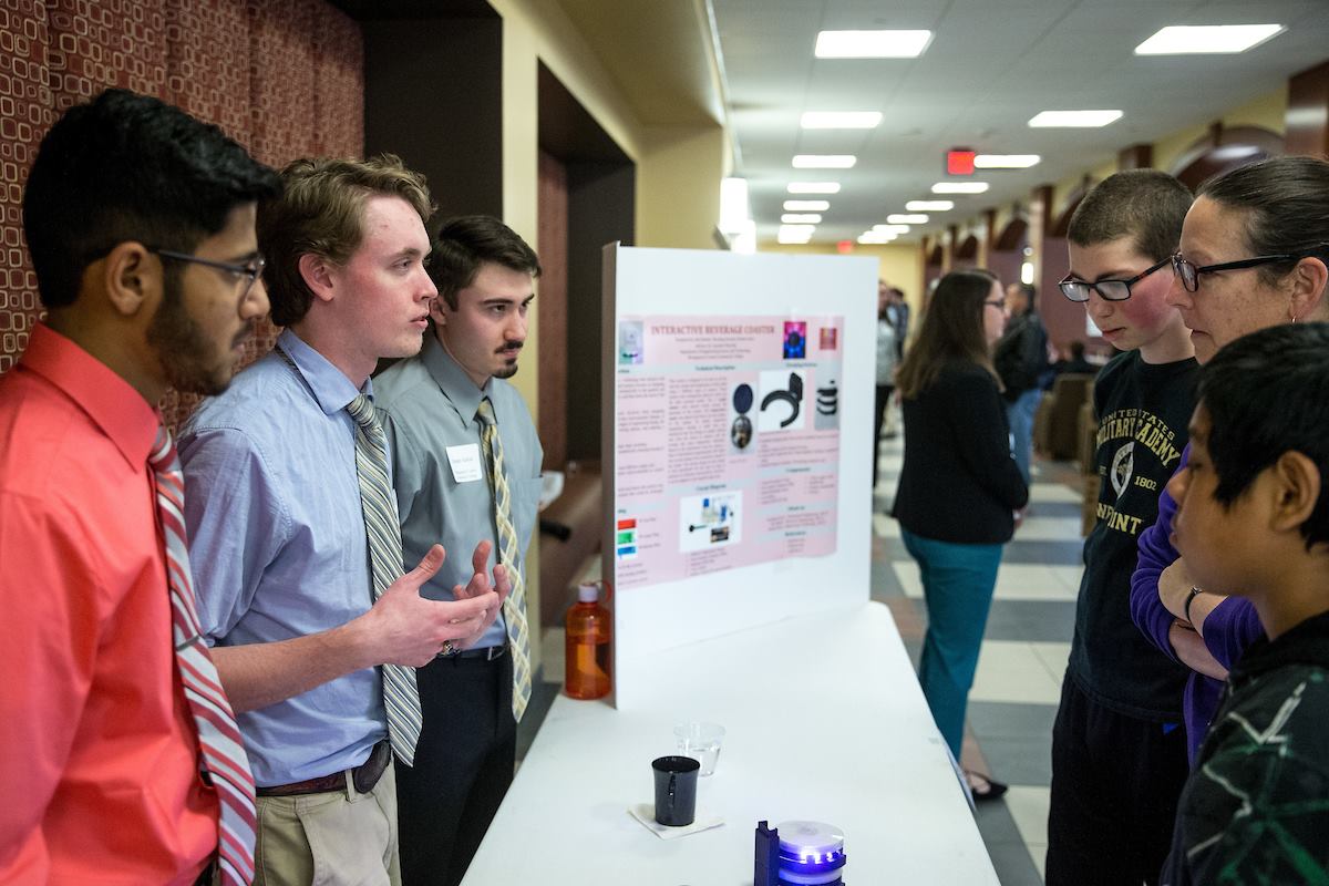 Three undergraduate researchers answer questions from onlookers about their researchers. The student on the left is wearing a red oxford shirt; the middle student is wearing a blue oxford shirt, and the student on the right is wearing a green oxford shirt. Their poster is in the center of photo. An adult with two children listen.