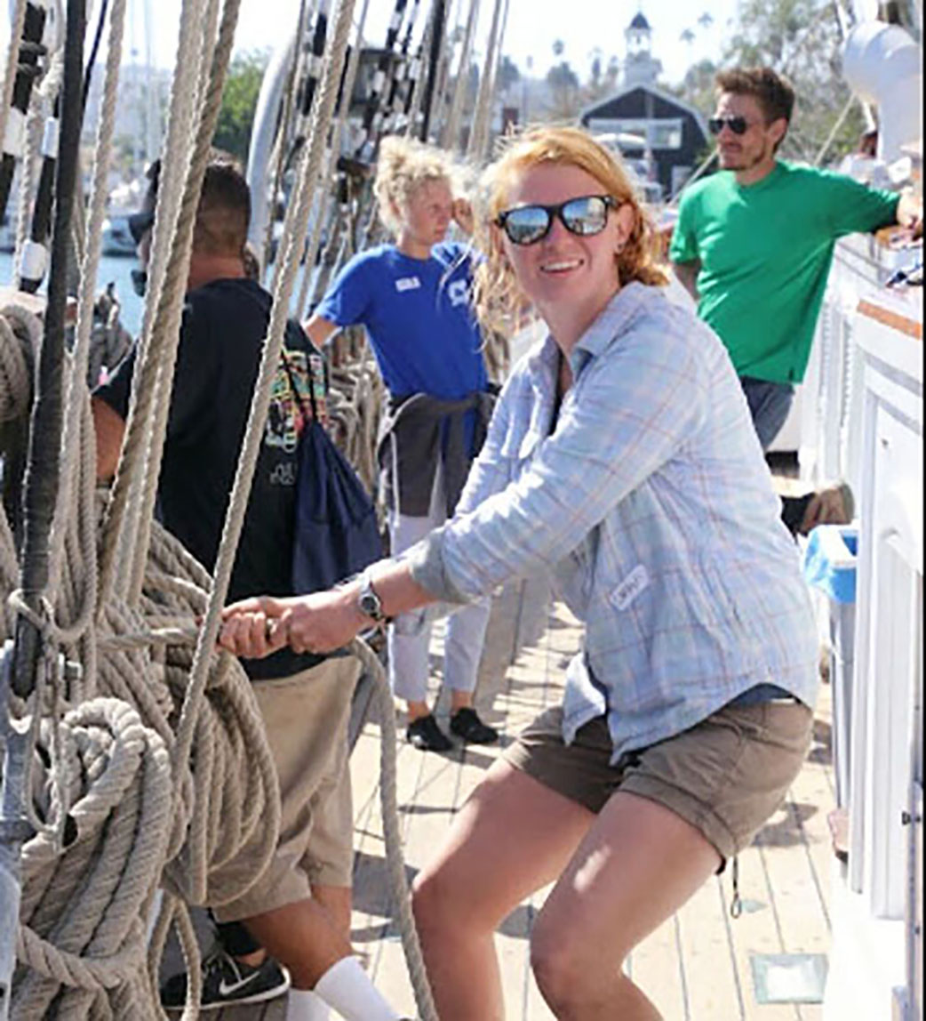 Carolyn Wasser on her boat, unraveling a rope and wearing sunglasses with other sailors in the background 