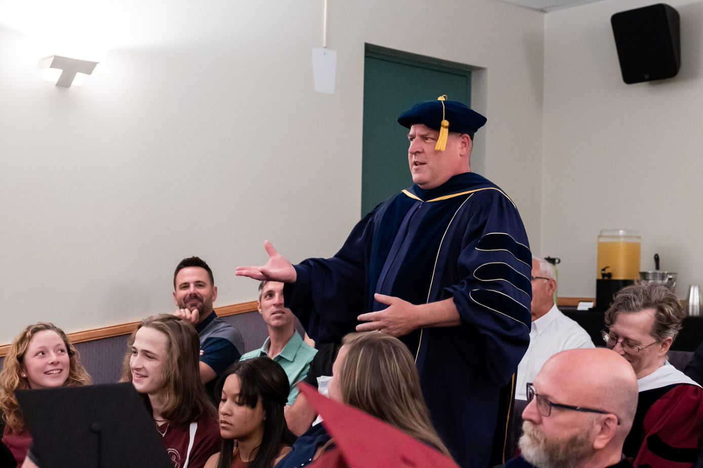Dr. Johnson wearing ceremonial robes, standing and presenting to a crowd of people who are seated around him 