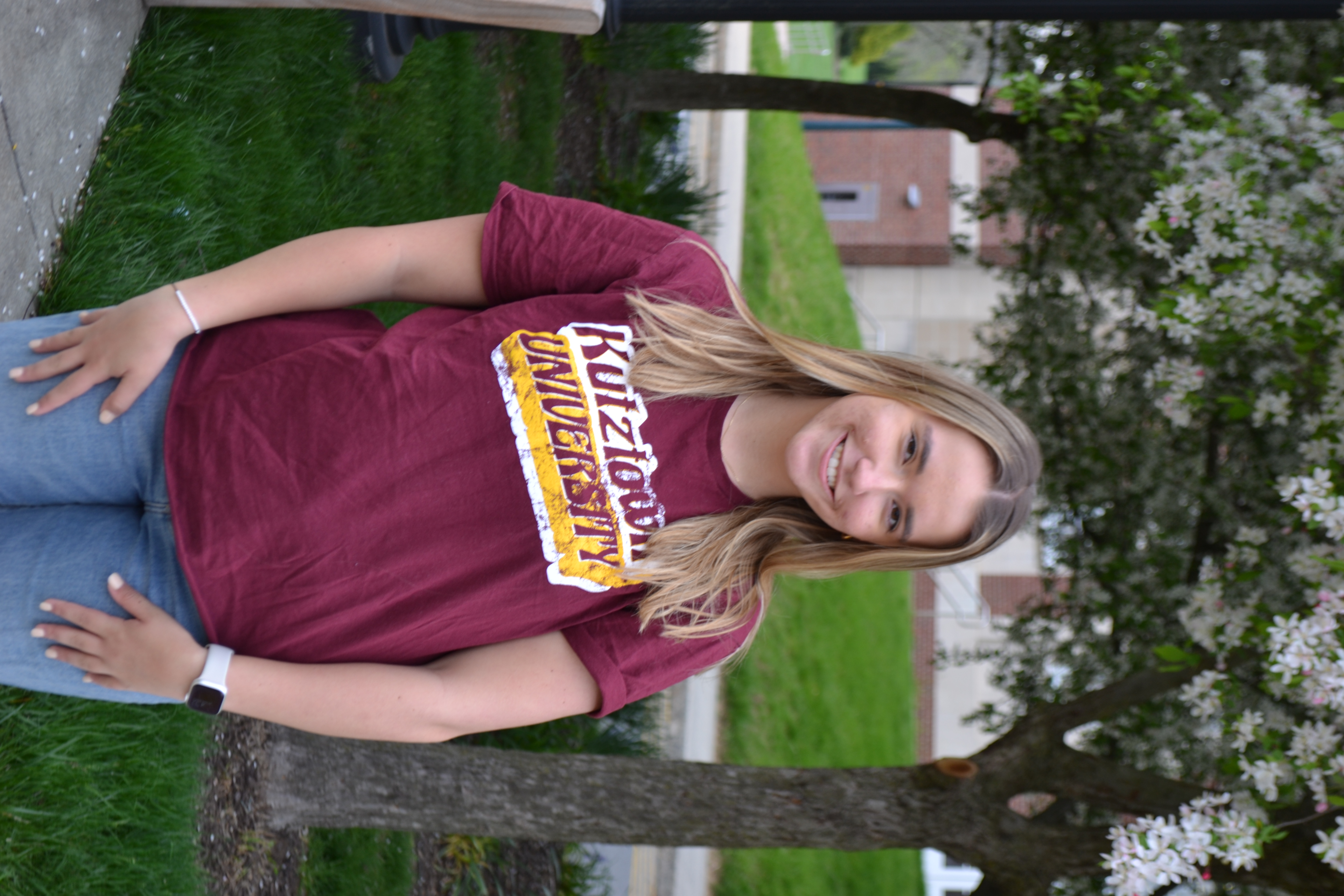 Transfer student Maegan wearing a maroon K-U t-shirt standing outside in front of trees on campus, smiling at the camera 