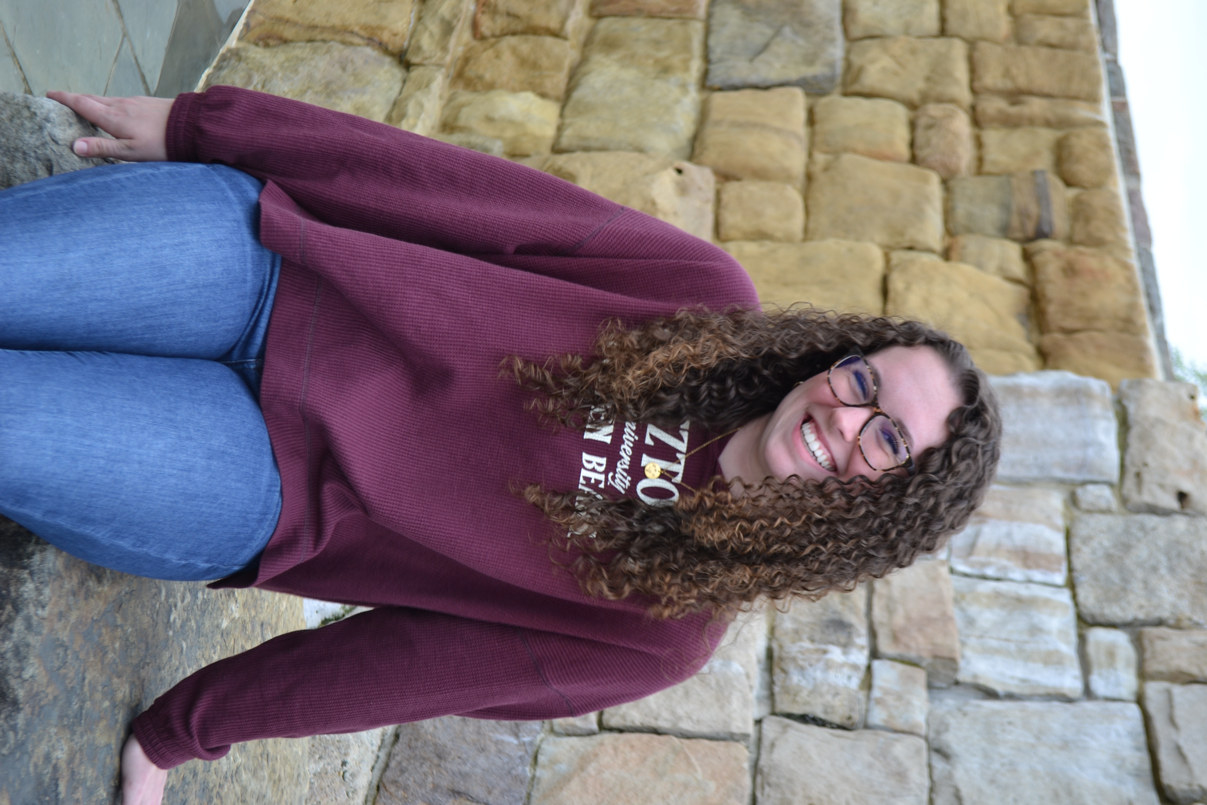 Transfer student Emma sitting on the Alumni Plaza Fountain wearing a K-U sweatshirt