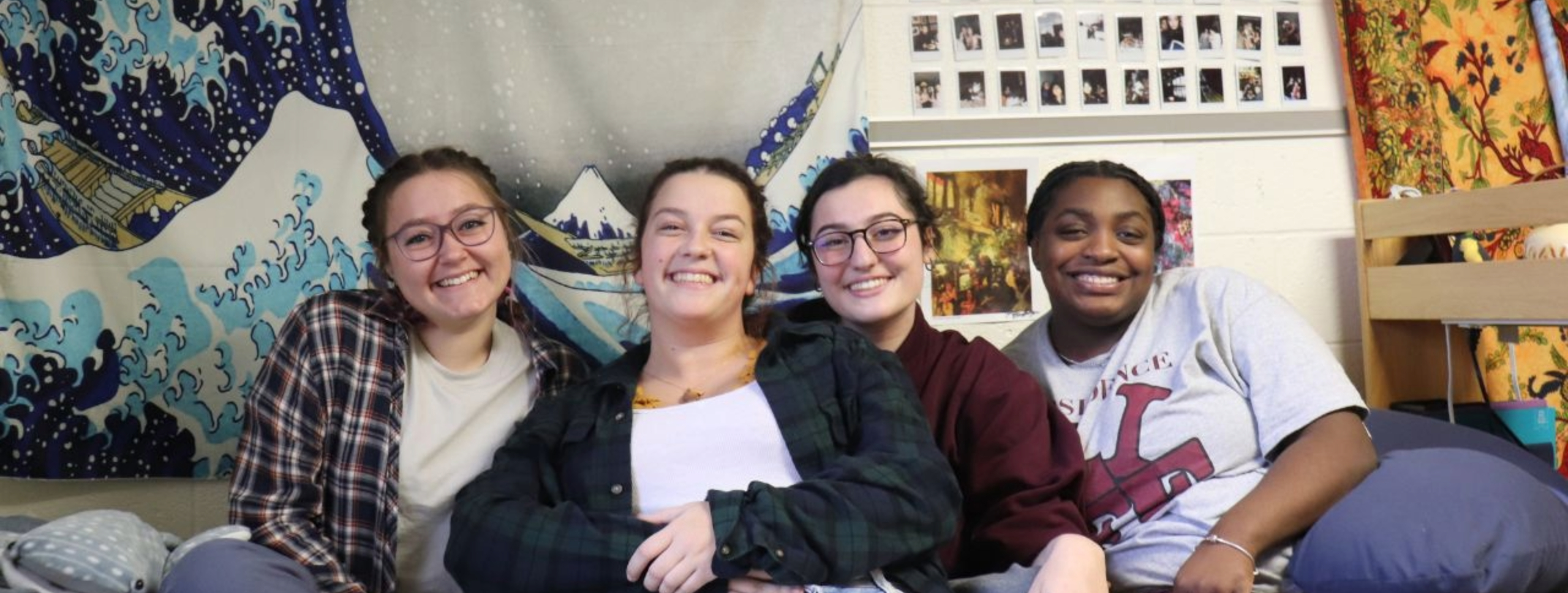four students sitting on a residence hall room bed smiling