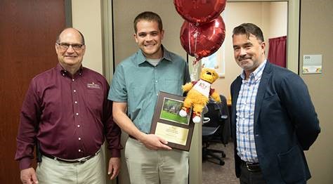 Jared Green holding his employee of the month placard, stuffed golden bear, and celebratory balloons, with members of the HR council standing on either side of him