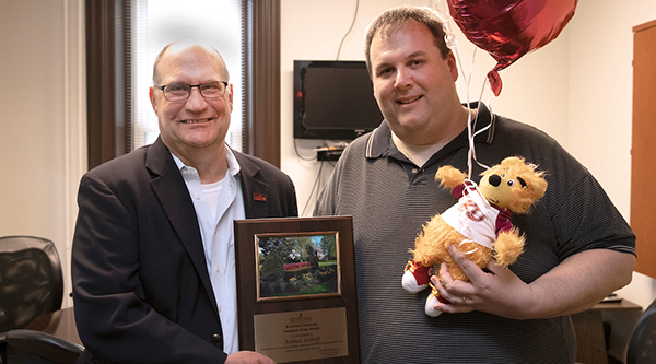 Dr. Hawkinson presenting Joshua Leiboff with the employee of the month placard; each man is holding one side of the award, and Leiboff also holds a stuffed golden bear and celebratory balloons 