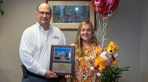 Christina Ferris holding her celebratory balloons and stuffed golden bear, while Dr. Hawkinson presents her with the Employee of the Month placard 