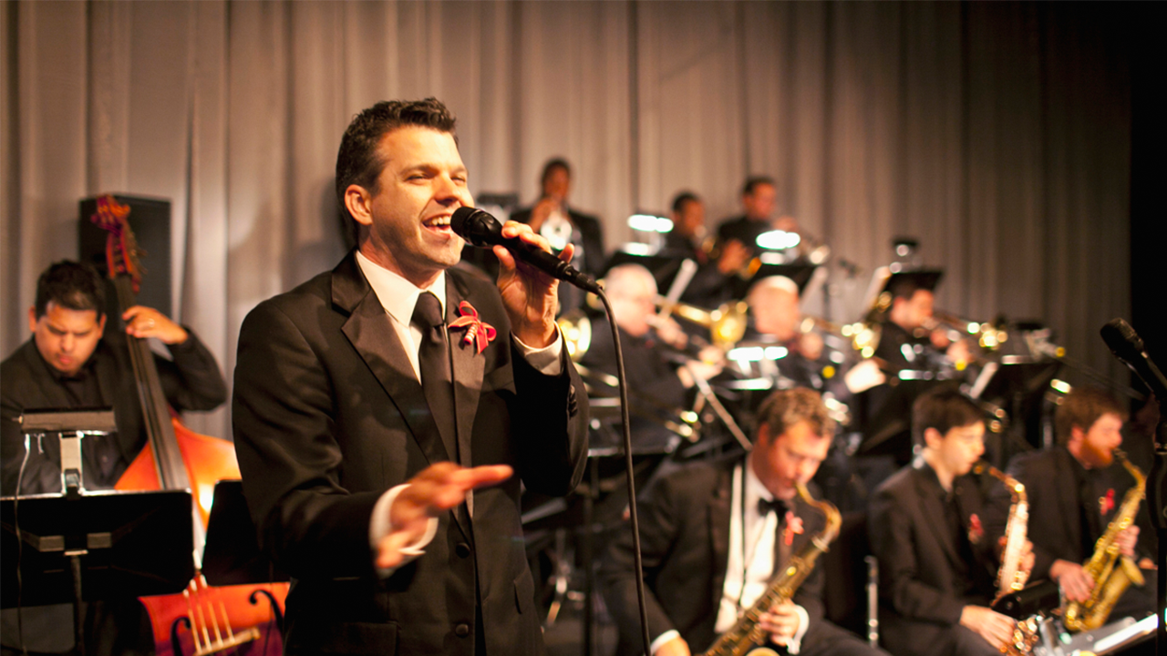 Male student in a tuxedo, singing on stage with the orchestra playing in the background